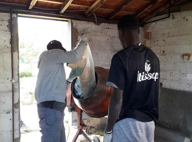 Deux hommes travaillant à l'intérieur d'un bâtiment, l'un versant un sac de mélange sec dans une bétonnière rouge.